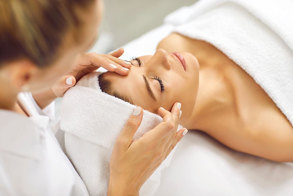 Spa therapist applying facial treatment to a relaxed client lying on a white towel in a bright spa room