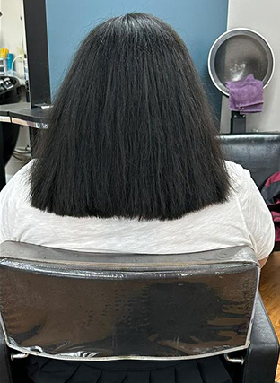 Person with straight black shoulder-length hair seated in a salon chair under a cape, back view
