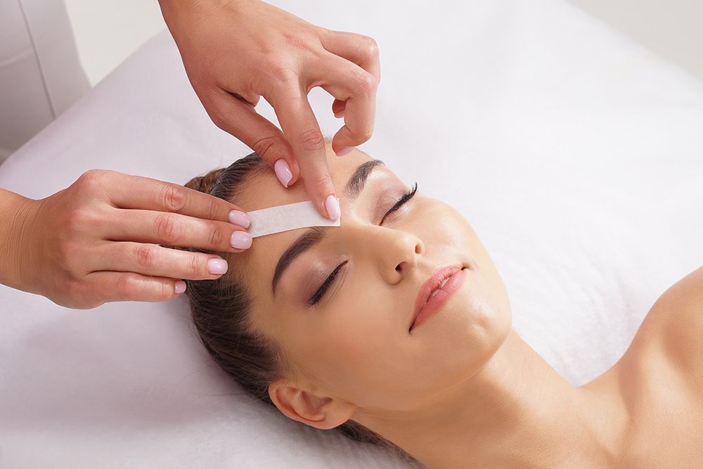 Hands apply a white eyebrow strip to a relaxed woman lying on a spa bed