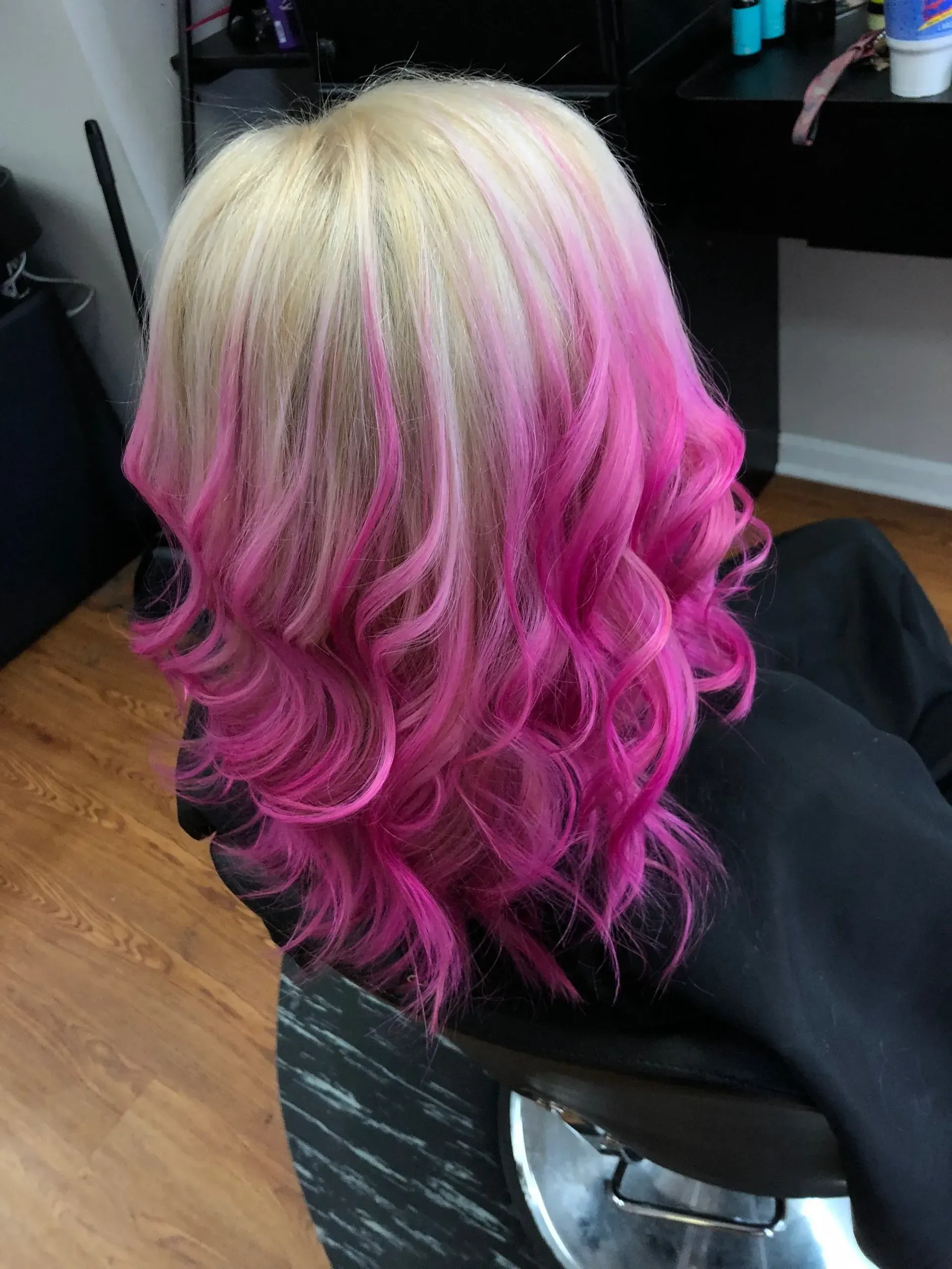 Short curly hairstyle with platinum blonde roots fading to bright pink ends in a salon chair