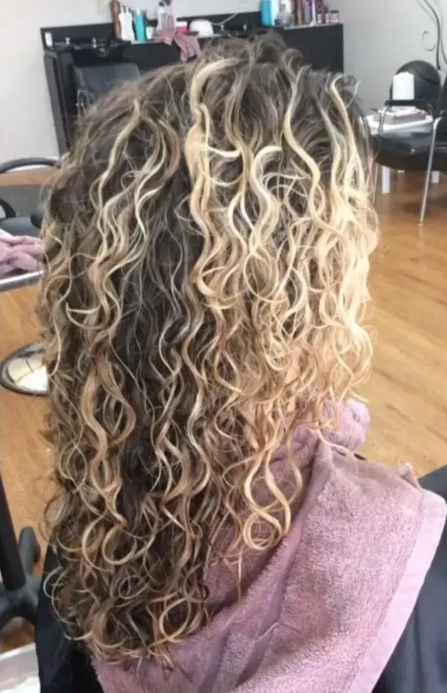 Curly highlighted hair from the back in a salon chair, with a purple cape draped over the shoulders.